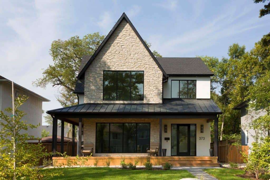 Modern two-story house with a gabled roof, featuring large black-framed windows, light-colored stone facade, and a covered front porch.